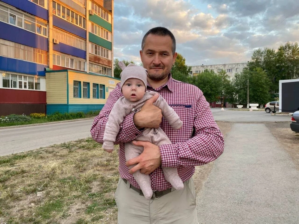 Тифлокомментарий: цветная фотография. Улица в городском квартале подсвечена теплым светом заходящего солнца. На асфальтированной дорожке, тянущейся вдоль многоэтажного жилого дома, стоит Руслан с младенцем на руках, с улыбкой прижимает его к своей груди. У Руслана очень короткие темные волосы с проседью у висков, овальное лицо с правильными чертами, карие глаза. На нем красно-белая клетчатая рубашка и бежевые брюки. На малыше бежевые комбинезон и шапочка с заячьими ушками. Тифлокомментарий: цветная фотография. Улица в городском квартале подсвечена теплым светом заходящего солнца. На асфальтированной дорожке, тянущейся вдоль многоэтажного жилого дома, стоит Руслан с младенцем на руках, с улыбкой прижимает его к своей груди. У Руслана очень короткие темные волосы с проседью у висков, овальное лицо с правильными чертами, карие глаза. На нем красно-белая клетчатая рубашка и бежевые брюки. На малыше бежевые комбинезон и шапочка с заячьими ушками.